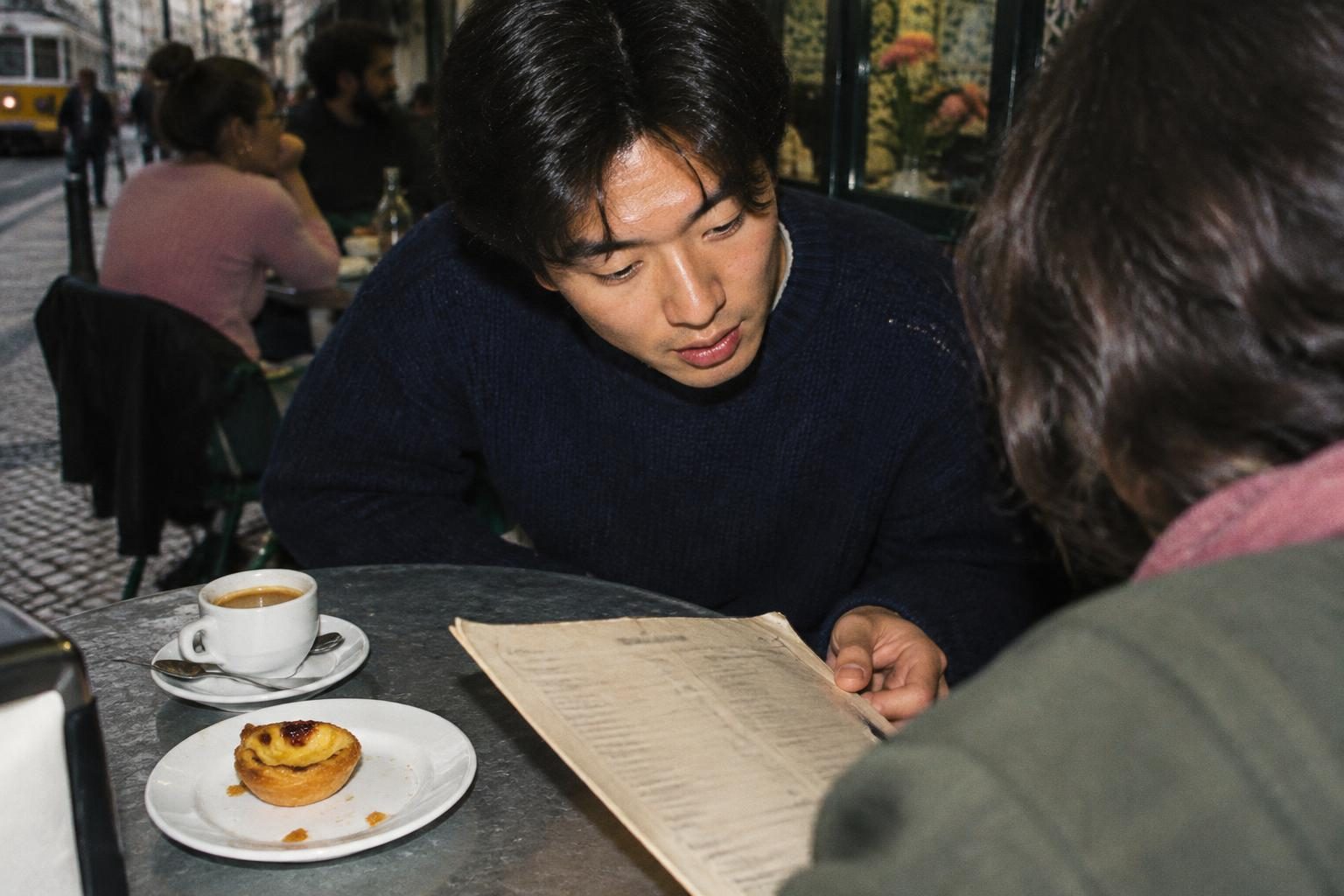 A traveller studies a worn menu over espresso and a custard pastry in Lisbon.